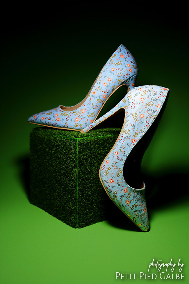 Blue high heels with flower pattern standing on a green grass cube prop, low key lighting.